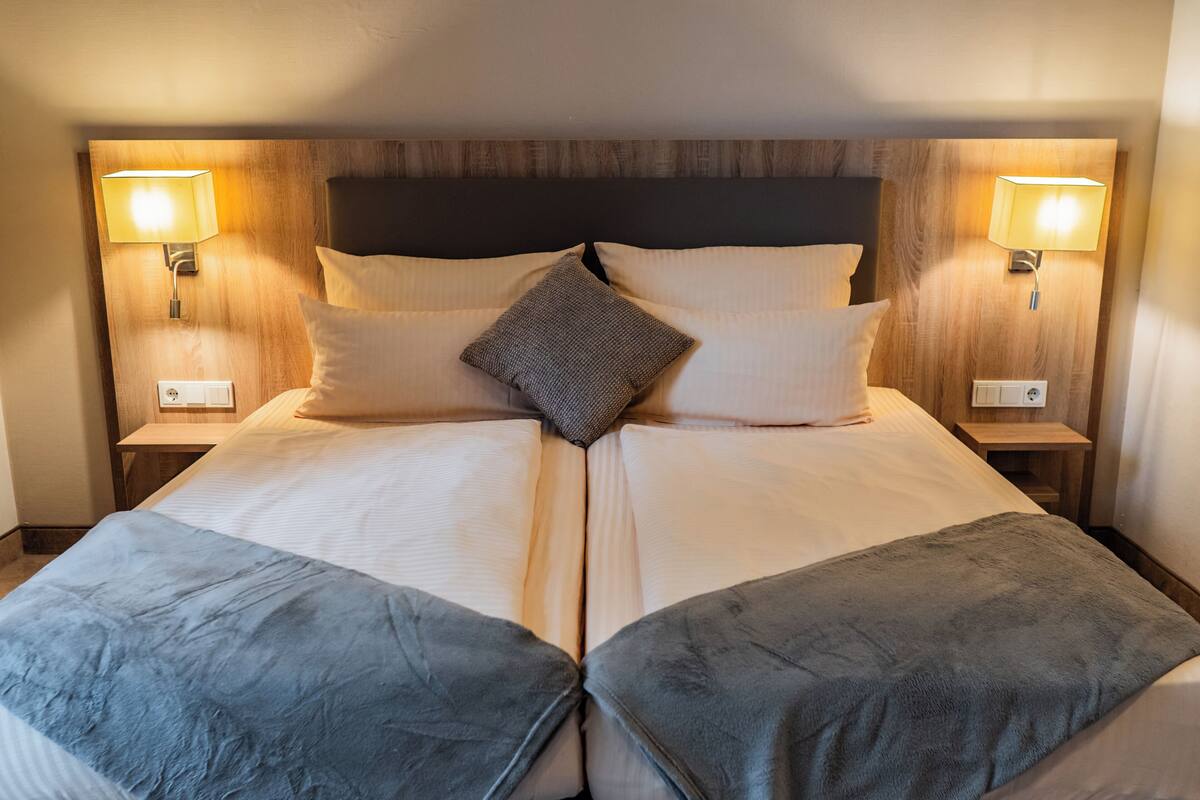 Una cama doble con cabecera de madera, dos lámparas de noche y un cojín decorativo central.