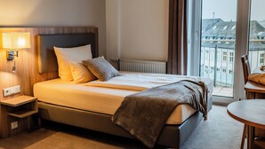 Habitación de hotel con cama, mesita de noche, lámpara y vista a la ciudad por una ventana.