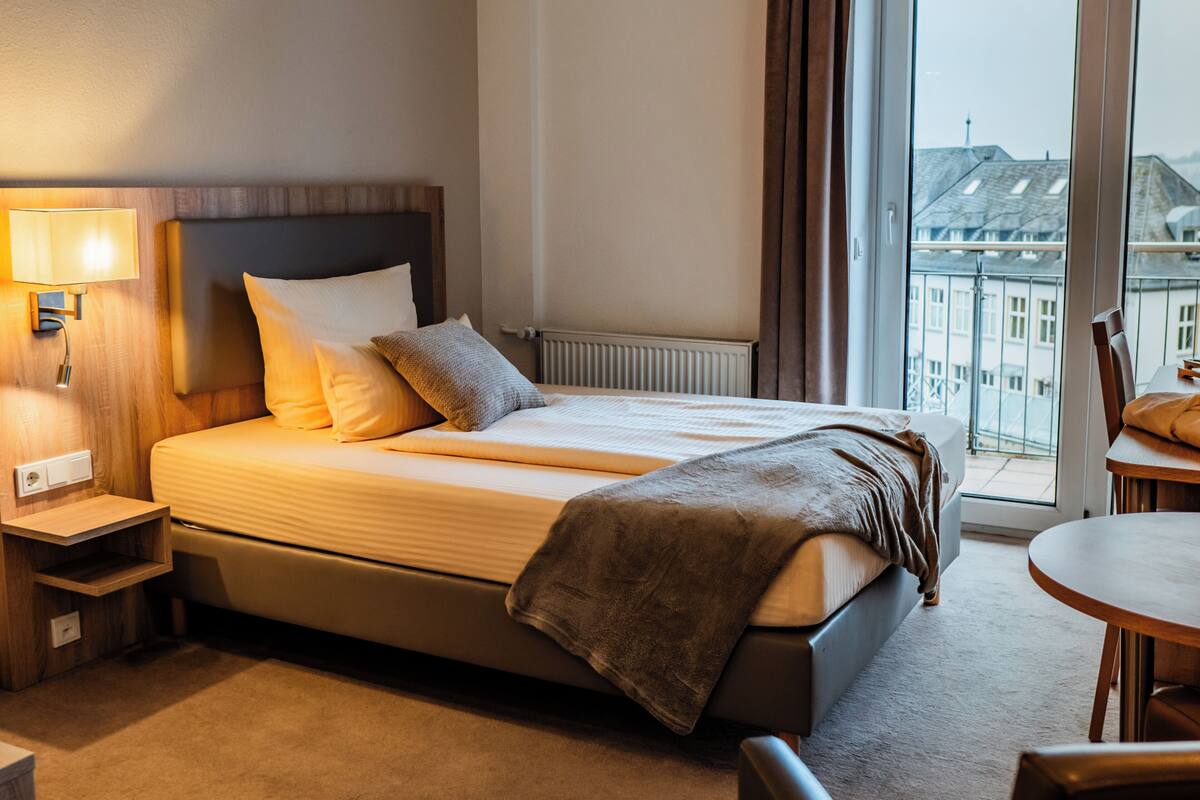Habitación de hotel con cama, mesita de noche, lámpara y vista a la ciudad por una ventana.
