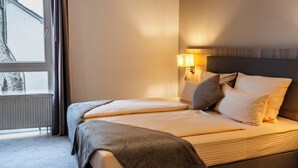 Una habitación de hotel con cama, mesita de noche, lámpara y ventana con cortinas.