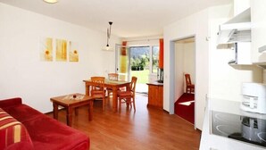 Un salon moderne avec un canapé rouge, une table à manger et des chaises en bois, un coin cuisine avec une cuisinière, et une vue sur l’extérieur grâce à une porte vitrée.