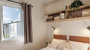 A bedroom with a bed, a window with curtains, a bedside lamp, and a shelf with books and a plant. - Camping Lanterna - Eurocamp (Tar)
