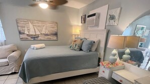 A bedroom with a bed, bedside tables, a chair, a bench, a ceiling fan, and artwork on the walls. - Under the Sea (Galveston)