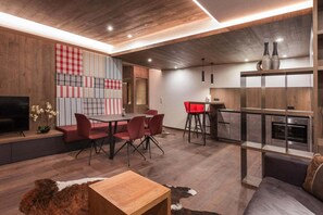 A modern living room with a wooden ceiling, a dining area with red chairs, a kitchen with a glass partition, and a TV on a wooden shelf. - Apartment in Ischgl with sauna (Ischgl)