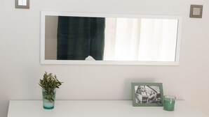 Un aparador blanco con espejo, un cuadro enmarco, una vela verde y un florero de vidrio con plantas.