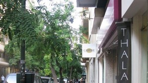 Una acera pavimentada con un diseño de ladrillo rojo, bordeada por coches estacionados y una cerca de metal negro. Un letrero de 'THALASSA COLLECTION' es visible a la derecha.