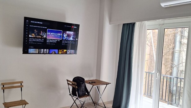 Una sala de estar moderna con un televisor de pantalla plana montado en la pared, una mesa plegable, una silla plegable, un banco y un ventanal con vistas al exterior.
