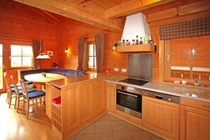 Interior de una cabaña de madera con cocina, que incluye estufa, horno y lavamanos. Hay una mesa de comedor con sillas y una ventana con cortinas.