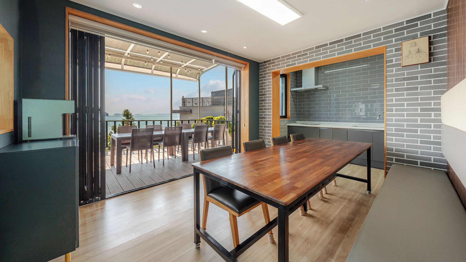 Una cocina moderna con salpicadero de ladrillo, un comedor con mesa y sillas de madera, y vista al mar a través de puertas correderas grandes.