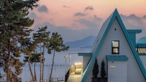 A building with a triangular roof, a staircase leading to a balcony, and a view of a body of water with trees in the foreground. - Sacheon Ara Private Pool Villa (Sacheon)