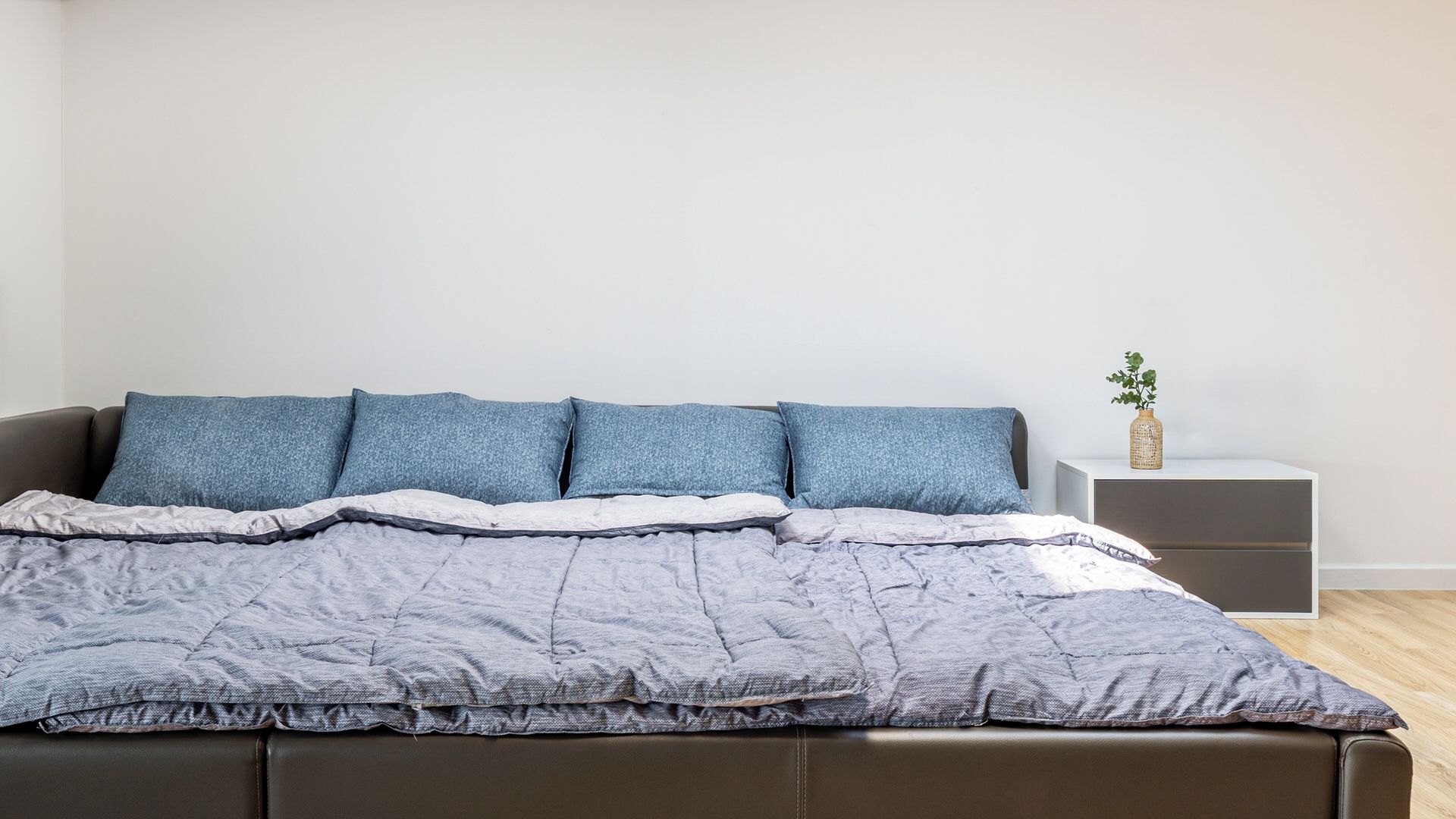 A bed with a grey quilt and four blue pillows, a bedside table with a vase, and a white nightstand.