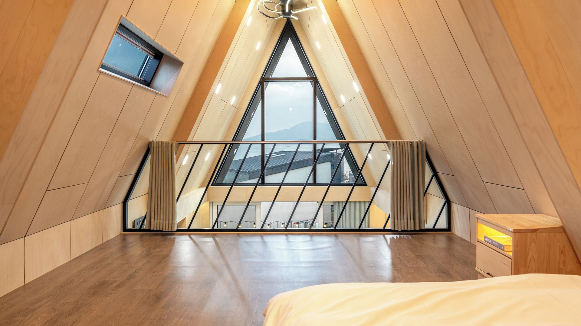 A room with a sloped ceiling, a bed, a small table, and a window with a view of a building.