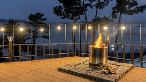 Eine Terrasse im Freien mit einem Metall-Feuerkorb, Lichterketten und Blick auf Bäume und Berge.