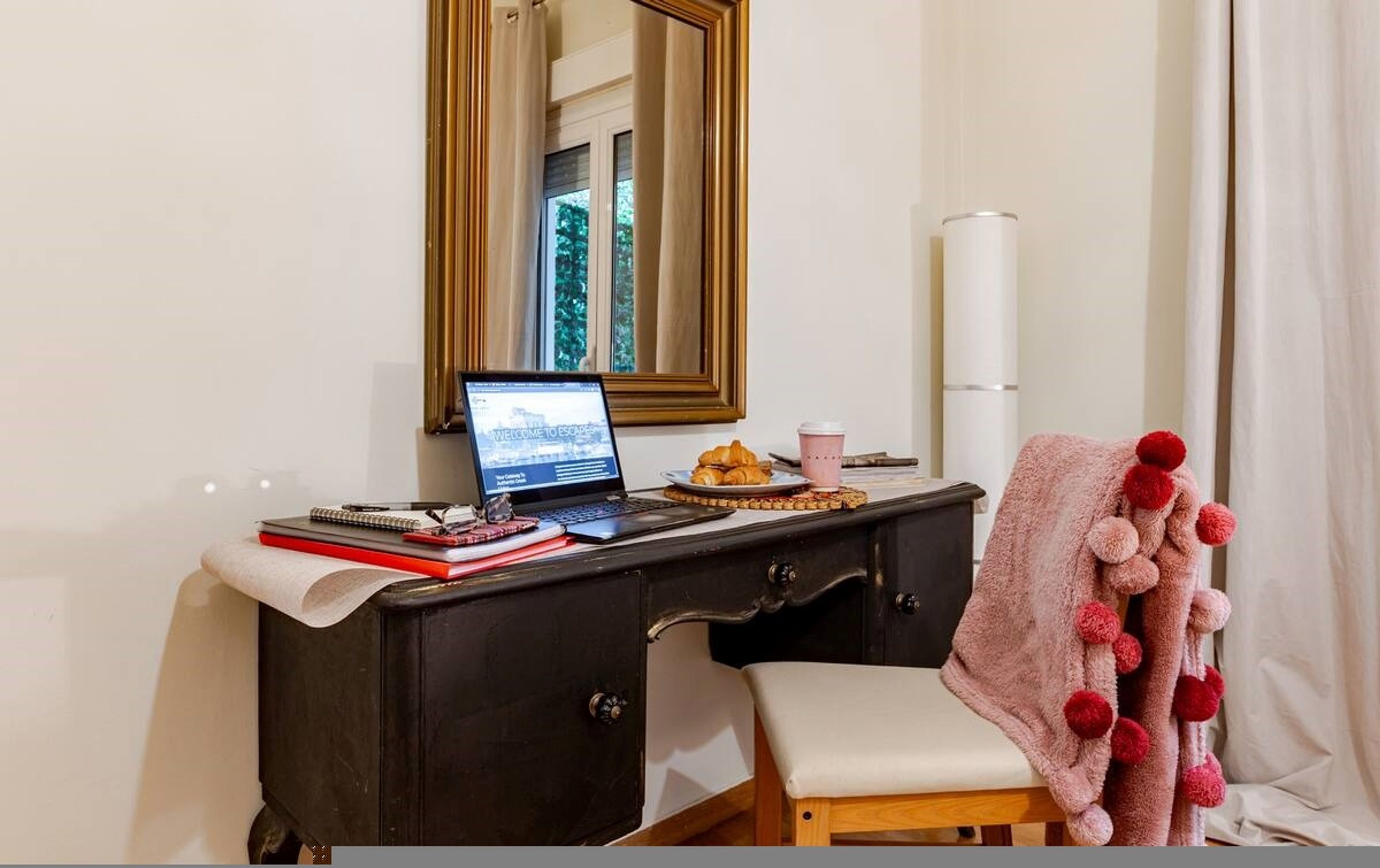 Ein Zimmer mit einem dunklen Holzkoffer, einem Laptop darauf, einem Teller mit Croissants, einer Kaffeetasse und einem Teddybären auf einem Stuhl.