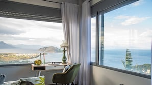 A room with a large window offering a view of a coastal landscape, a desk with a lamp, a chair, and decorative pillows. - La Casa del Sol Sea View Sunset Jacuzzi (Aegina)