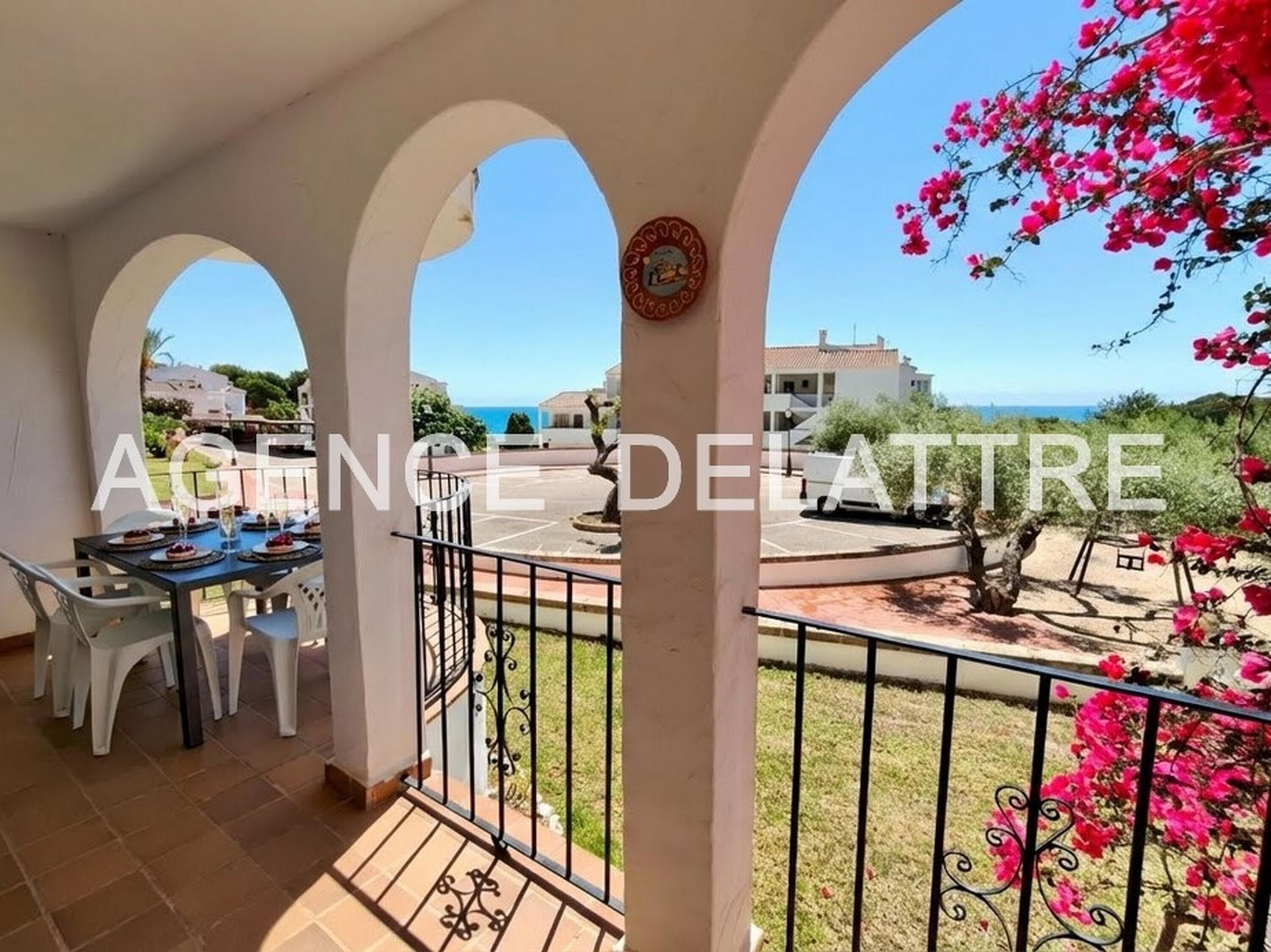 A balcony with a table set for dining, a view of the sea, and a garden with pink flowers.