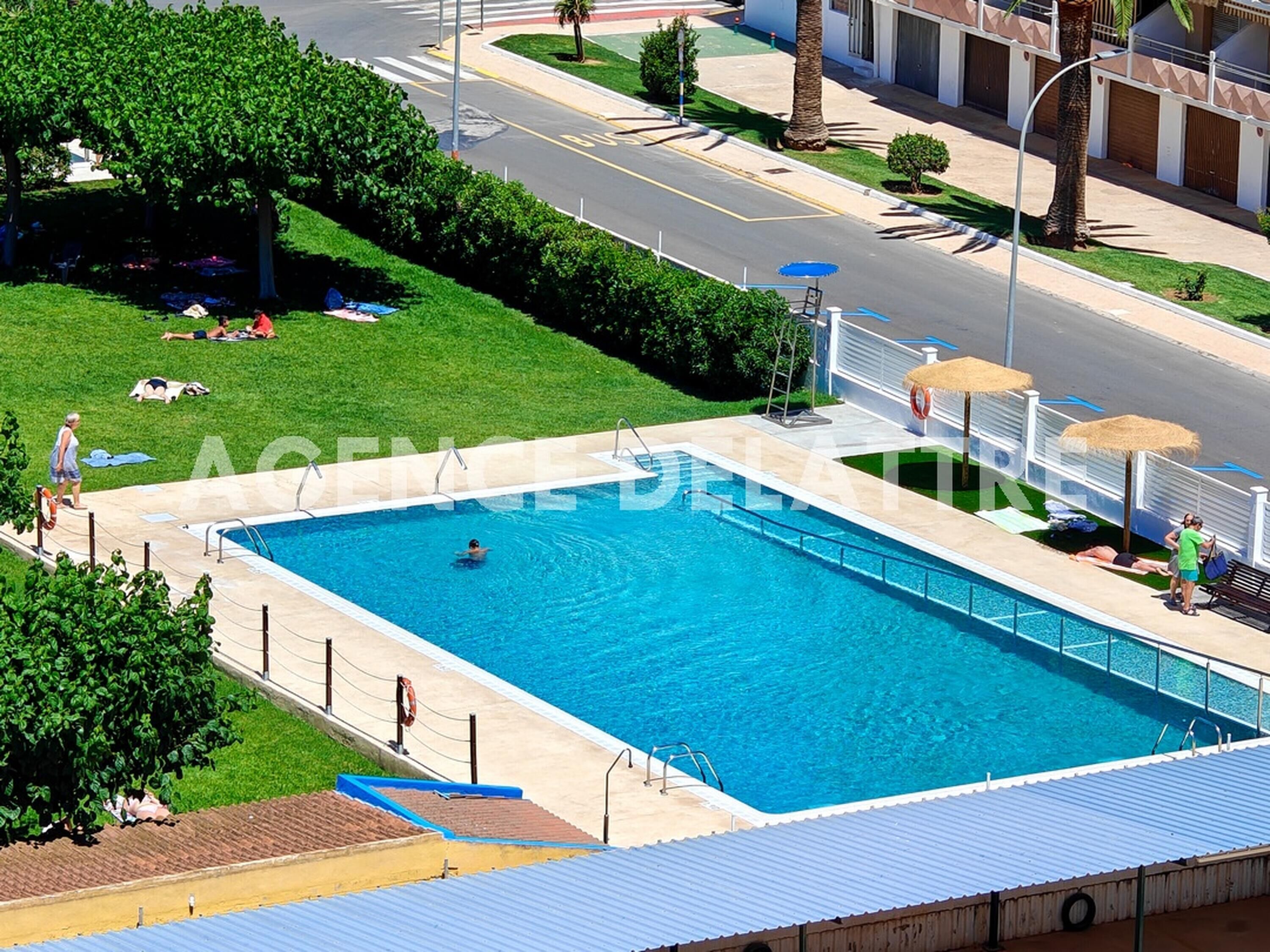 Une piscine à l’eau d’un bleu limpide, entourée d’une zone délimitée par une clôture, et située à côté d’une route bordée d’un trottoir et de verdure.
