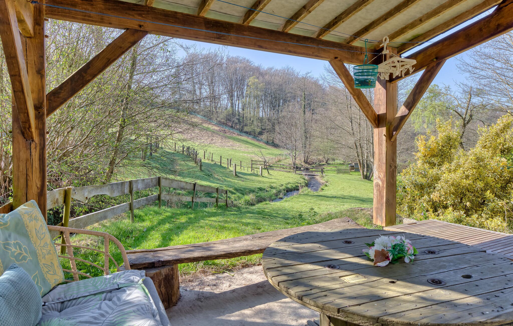 Une pergola en bois avec une table et des chaises, donnant sur un paysage verdoyant, agrémenté d’une clôture et d’arbres.