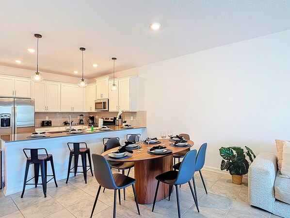Una cocina moderna con armarios blancos, una isla central con asientos y una zona de comedor con sillas y una mesa dispuesta para cuatro.