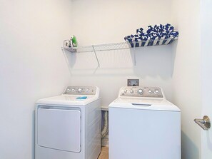 A laundry area with two white front-loading washing machines and a metal shelf above holding folded clothes. - Next to Epic Univers | Clubhouse Access Included (Davenport)