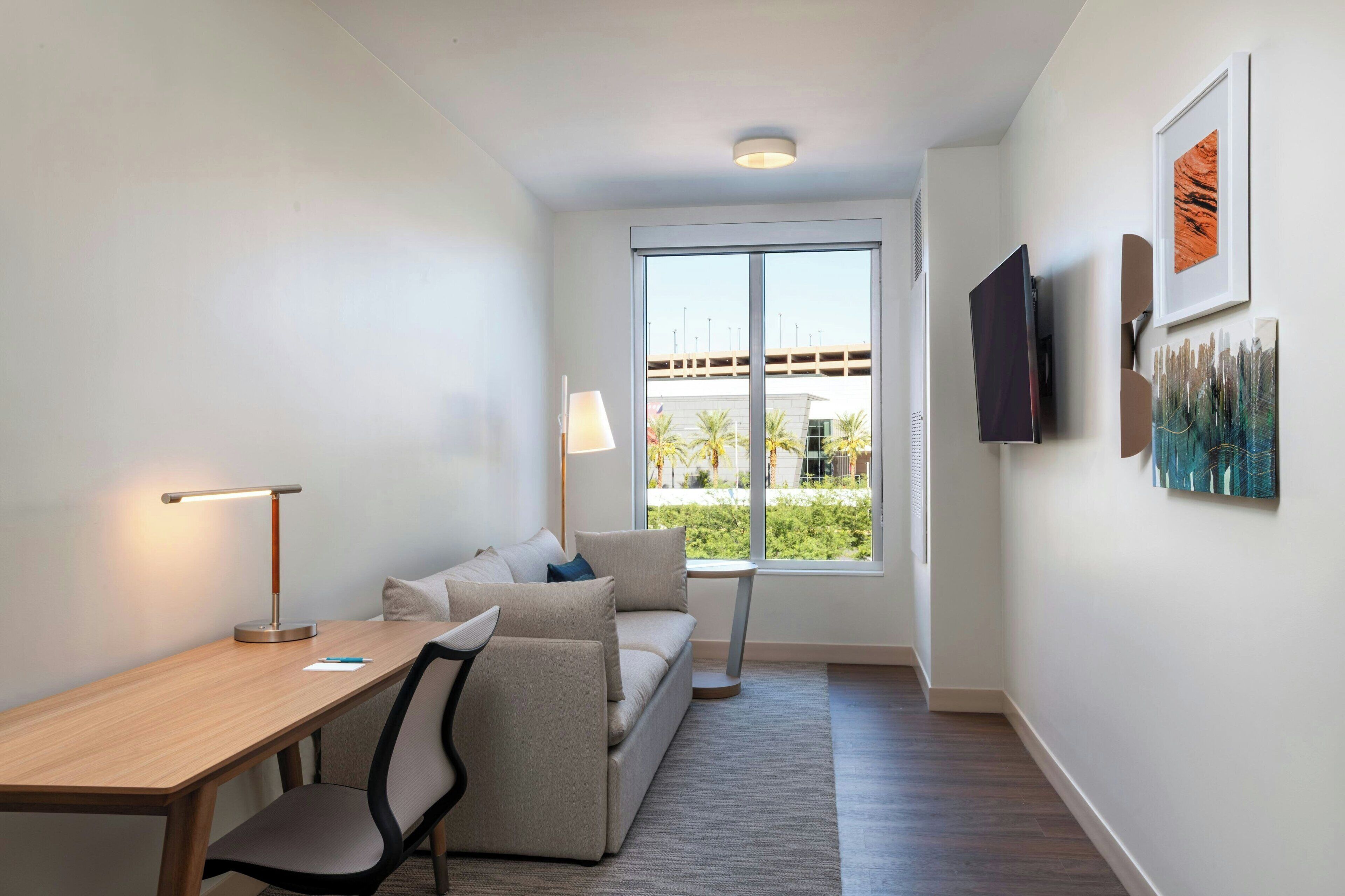 Une chambre d’hôtel moderne avec un bureau en bois, un canapé, un téléviseur fixé au mur et une grande fenêtre donnant sur des palmiers et un immeuble.