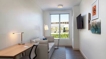 Une chambre d’hôtel moderne avec un bureau en bois, un canapé, un téléviseur fixé au mur et une grande fenêtre donnant sur des palmiers et un immeuble.