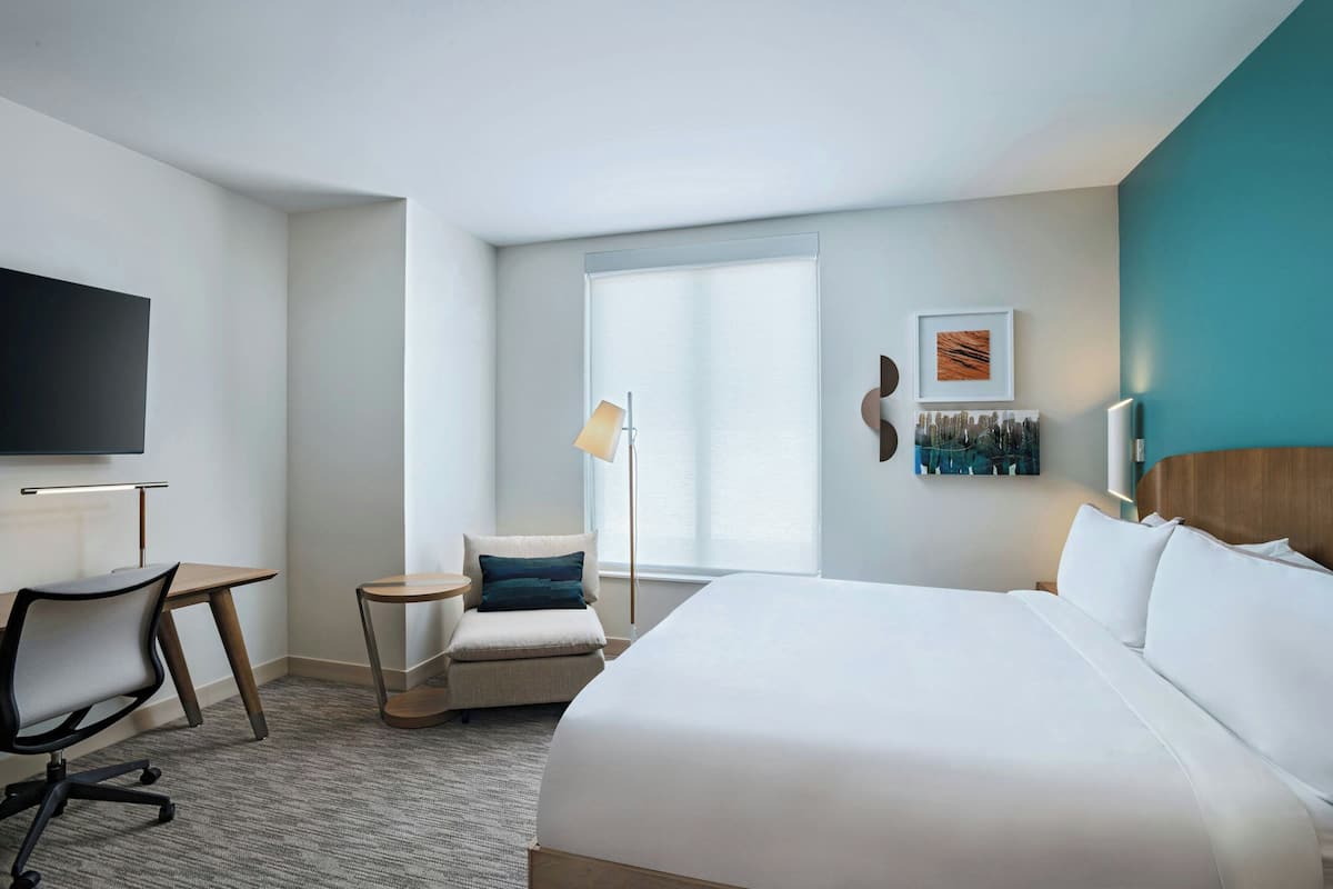 A hotel room with a large bed, a desk, a chair, a TV, and a window with blinds.