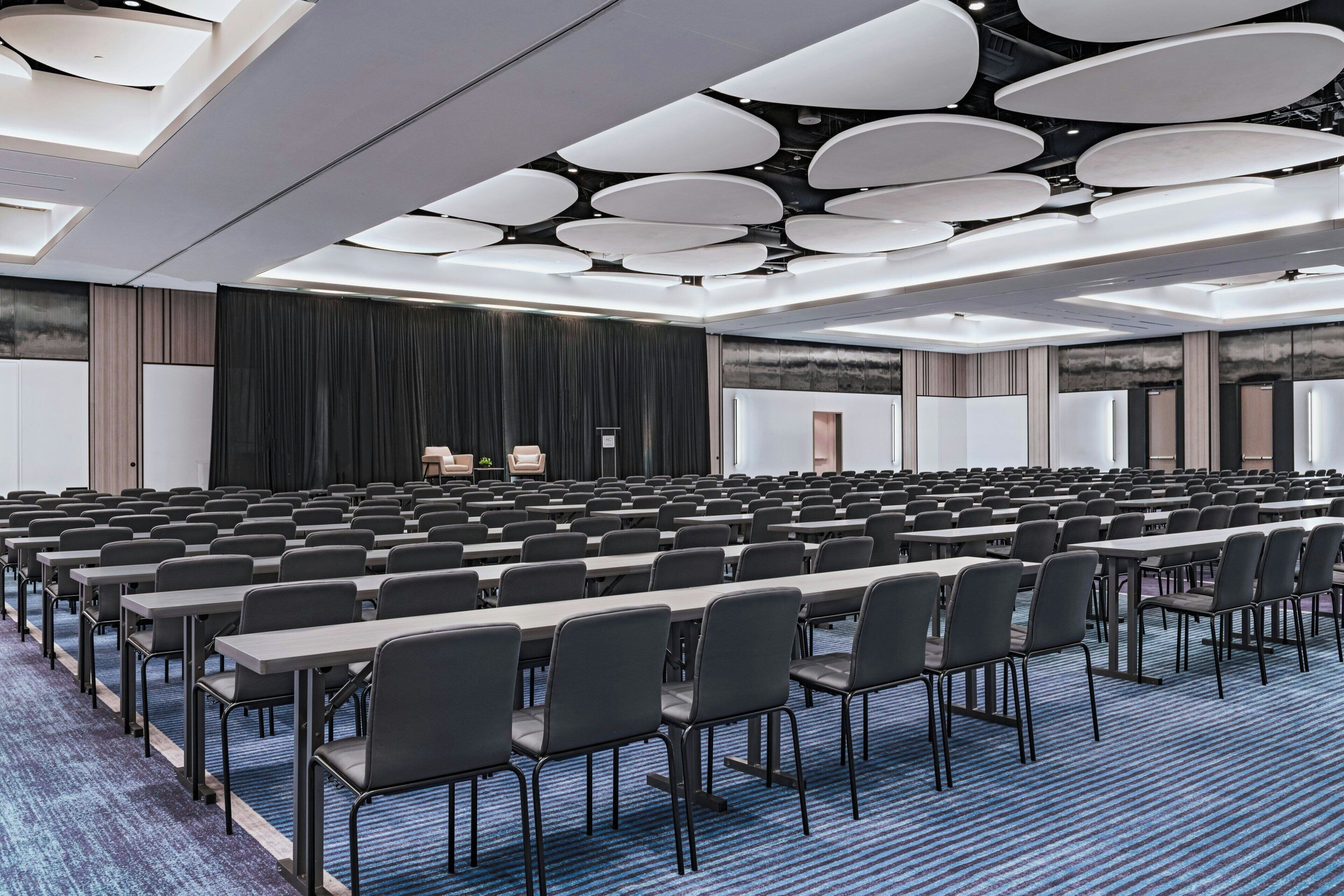 Ein Konferenzraum mit Tischen und Stühlen, einer Bühne und einem modernen Deckenstil.