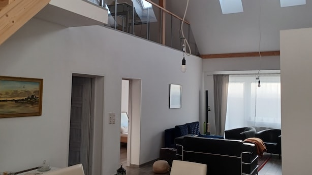 Un loft spacieux avec un plafond haut, des poutres apparentes, un coin repas avec une table et des chaises, un salon avec un canapé et une télévision, et un escalier menant à un niveau supérieur.