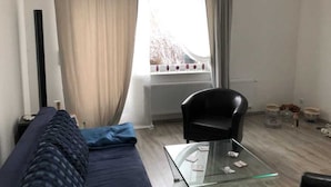 Une chambre d’hôtel avec un lit, une table basse en verre, un fauteuil noir, une fenêtre avec des rideaux et un radiateur.