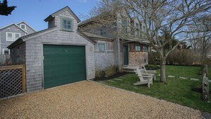 A two-story house with a green garage door, a small porch with chairs, and a tree in the yard. - Charming 3-Bedroom Home in the Heart of Oak Bluffs (Oak Bluffs)