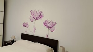 A bedroom with a bed, a nightstand, a lamp, and a wall with a floral mural. - At the home of "Nuccia" (Foglizzo)