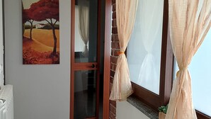 A hallway with a door leading to a room, a radiator, a painting of a landscape, and a window with curtains. - At the home of "Nuccia" (Foglizzo)