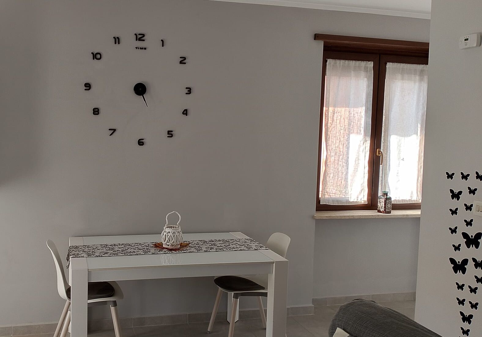 Un coin repas avec une table et des chaises blanches, une horloge murale, une fenêtre avec des rideaux et des autocollants représentant des papillons sur le mur.