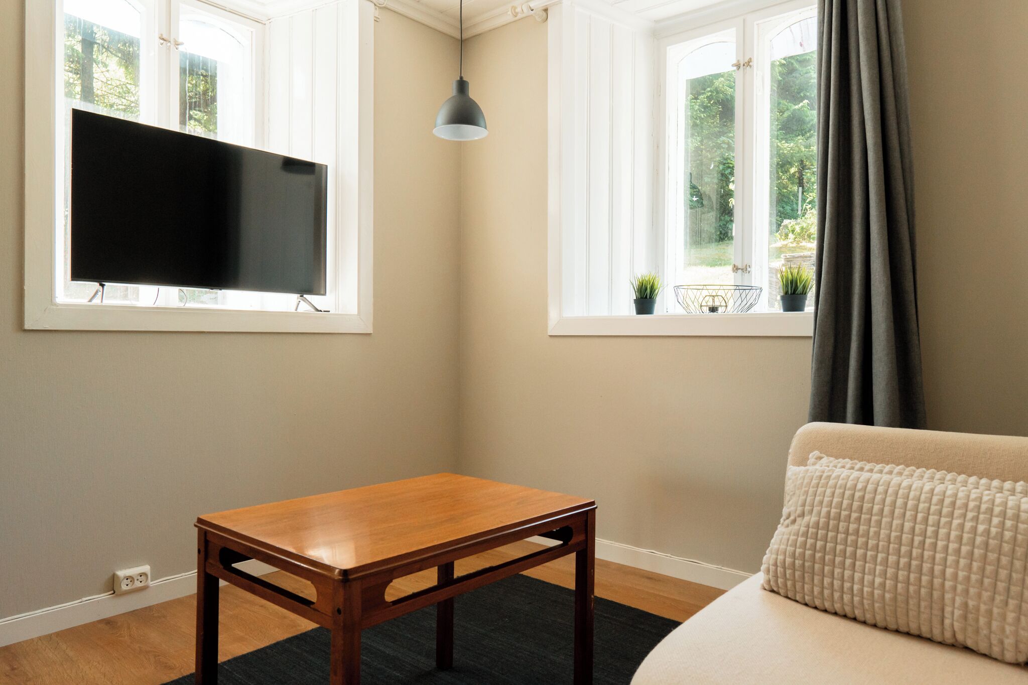 Una habitación con una mesa de madera, un sofá, un televisor y dos ventanas con cortinas.