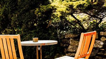 Dos sillas de madera y una pequeña mesa redonda en un entorno exterior con un muro de piedra y abundante vegetación al fondo.