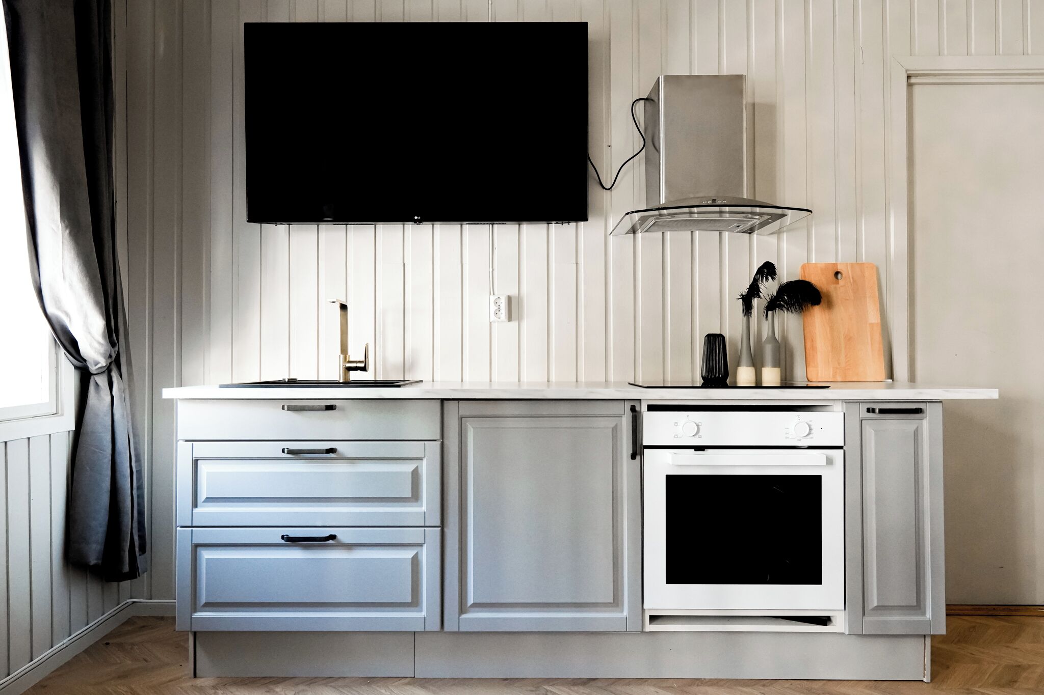 A kitchen with white cabinetry, a built-in oven, a microwave, a flat-screen TV mounted above the counter, and a wooden cutting board with utensils.