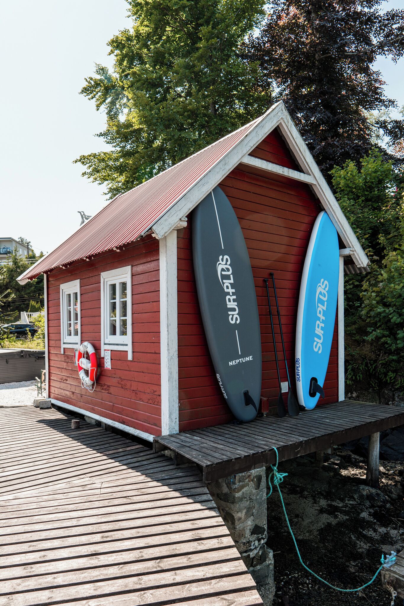 Een klein houten gebouw met twee surfplanken die ertegenaan leunen, een reddingsboei hangt aan de muur, en een houten terras ervoor.