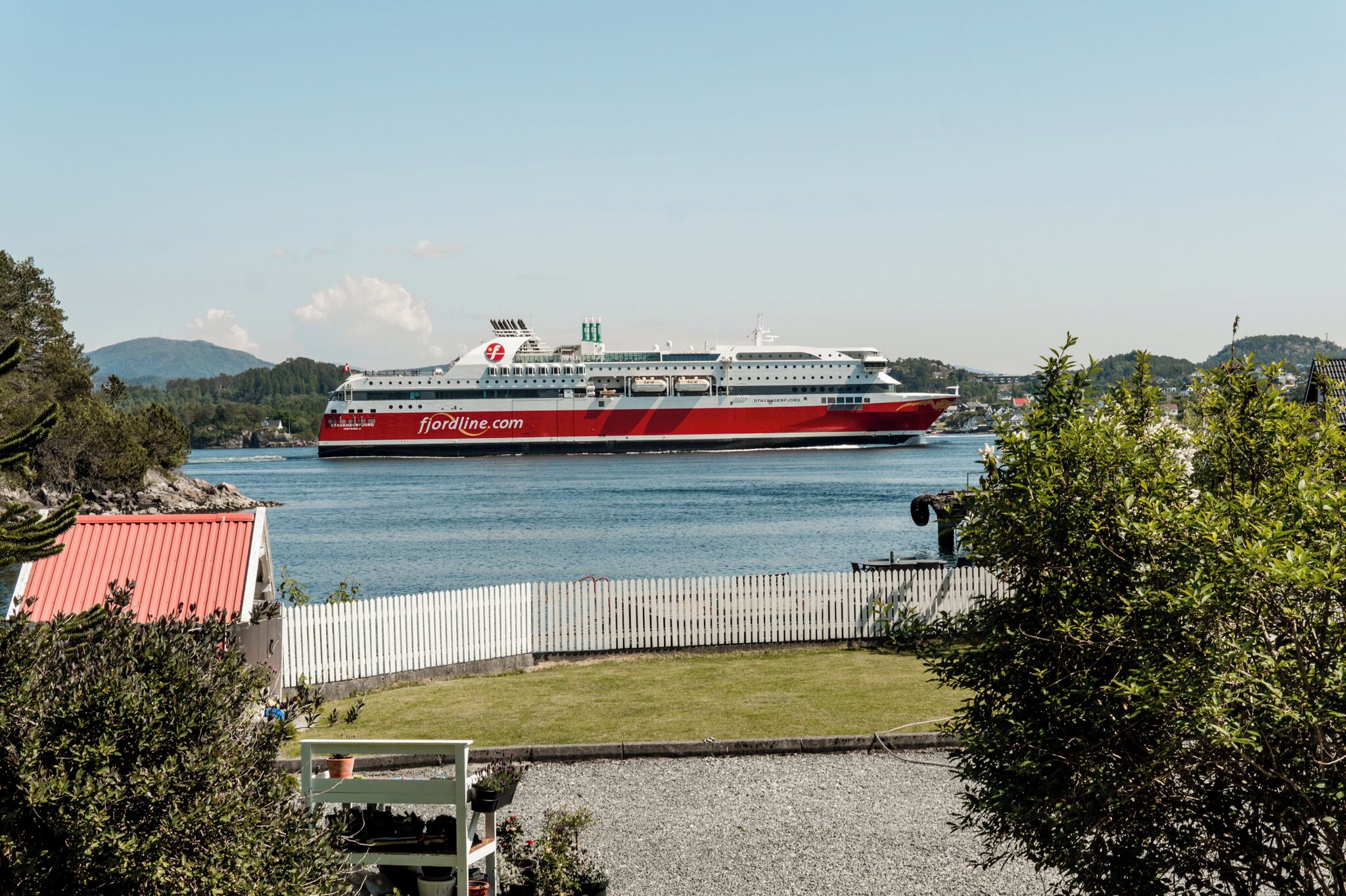 A large cruise ship docked at a port with a clear view of the surrounding landscape, including hills and a fenced area with greenery.