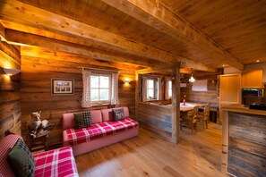 A cozy cabin interior with a wooden ceiling, a plaid sofa, a dining table, and a kitchen area.