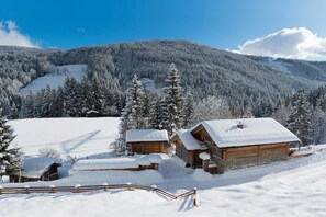 A wooden cabin with a snow-covered roof, surrounded by a wooden fence and snow-covered trees in a mountainous area.