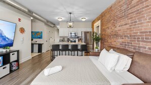 A modern hotel room with a large bed, a kitchen area, and a brick accent wall. - Steps Away From Mass Ave in Heart of Indy with Balcony (Indianapolis)
