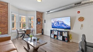 A living room with a TV, a dartboard, a bookshelf, and a dining area with chairs and a table. - Steps Away From Mass Ave in Heart of Indy with Balcony (Indianapolis)