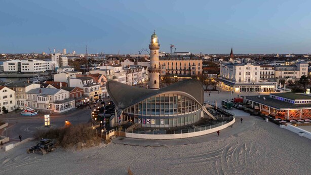 Een kuststadsgezicht met een opvallende toren, een modern gebouw met een gebogen dak en een promenade aan het water.