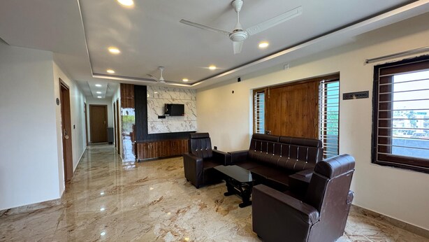 A modern living room with a marble floor, leather sofa, ceiling fan, and a TV mounted on the wall. - HOTEL MADHAVA RESIDENCY (Kakinada)