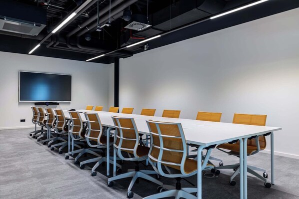 A conference room with a long table, multiple chairs, a flat-screen TV, and a modern ceiling with exposed ductwork. - NOTO Hotel (Reckange-sur-Mess)