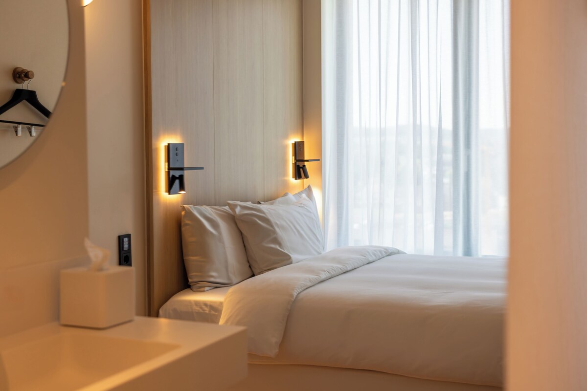 Une chambre d’hôtel avec un lit, des tables de chevet, une lampe fixée au mur et une fenêtre avec des rideaux.