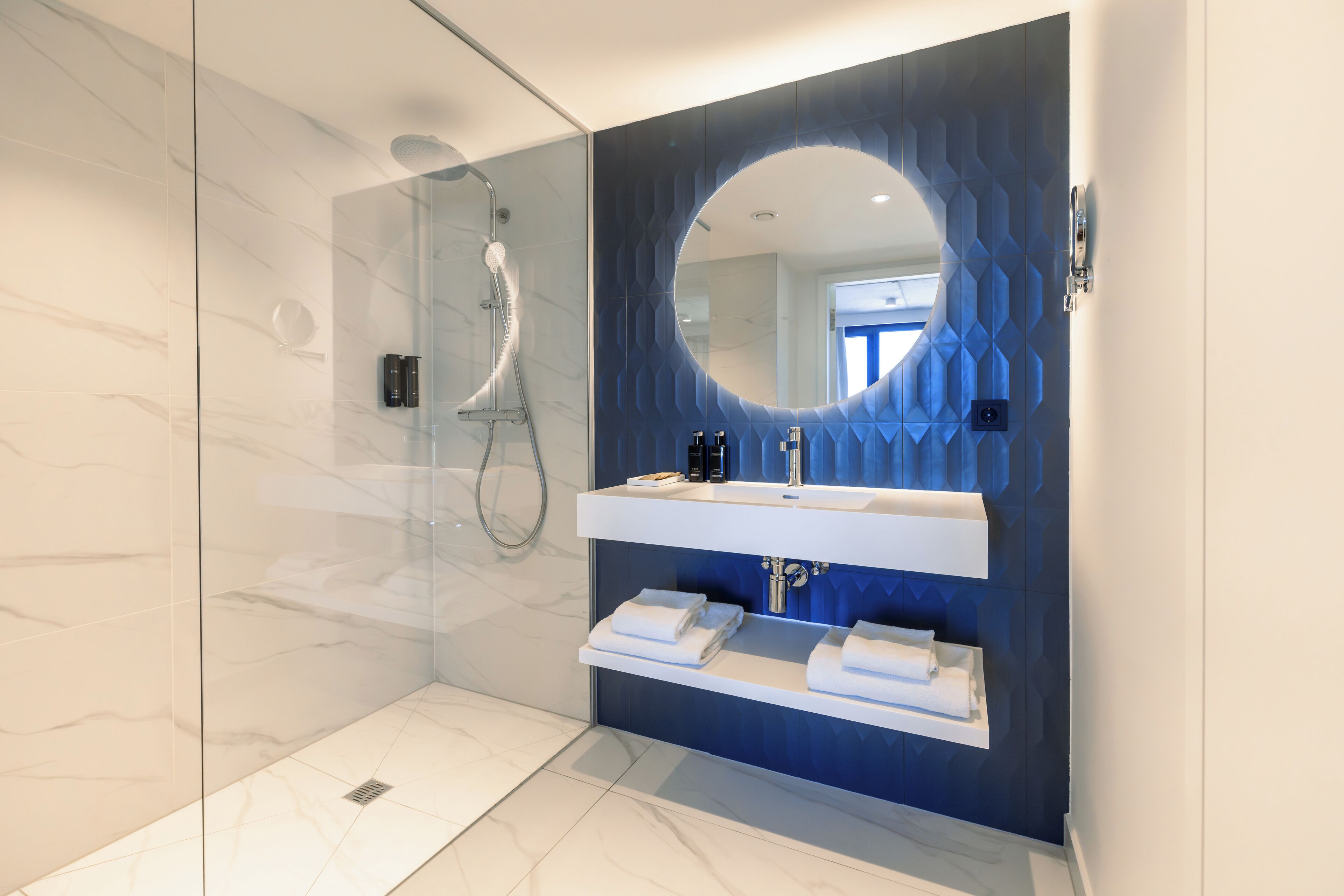 A modern bathroom with a glass-enclosed shower, a white vanity with a blue geometric backsplash, and neatly folded towels on a shelf.