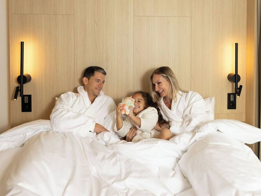 A family of four in a hotel room with a wooden headboard and bedside lamps.