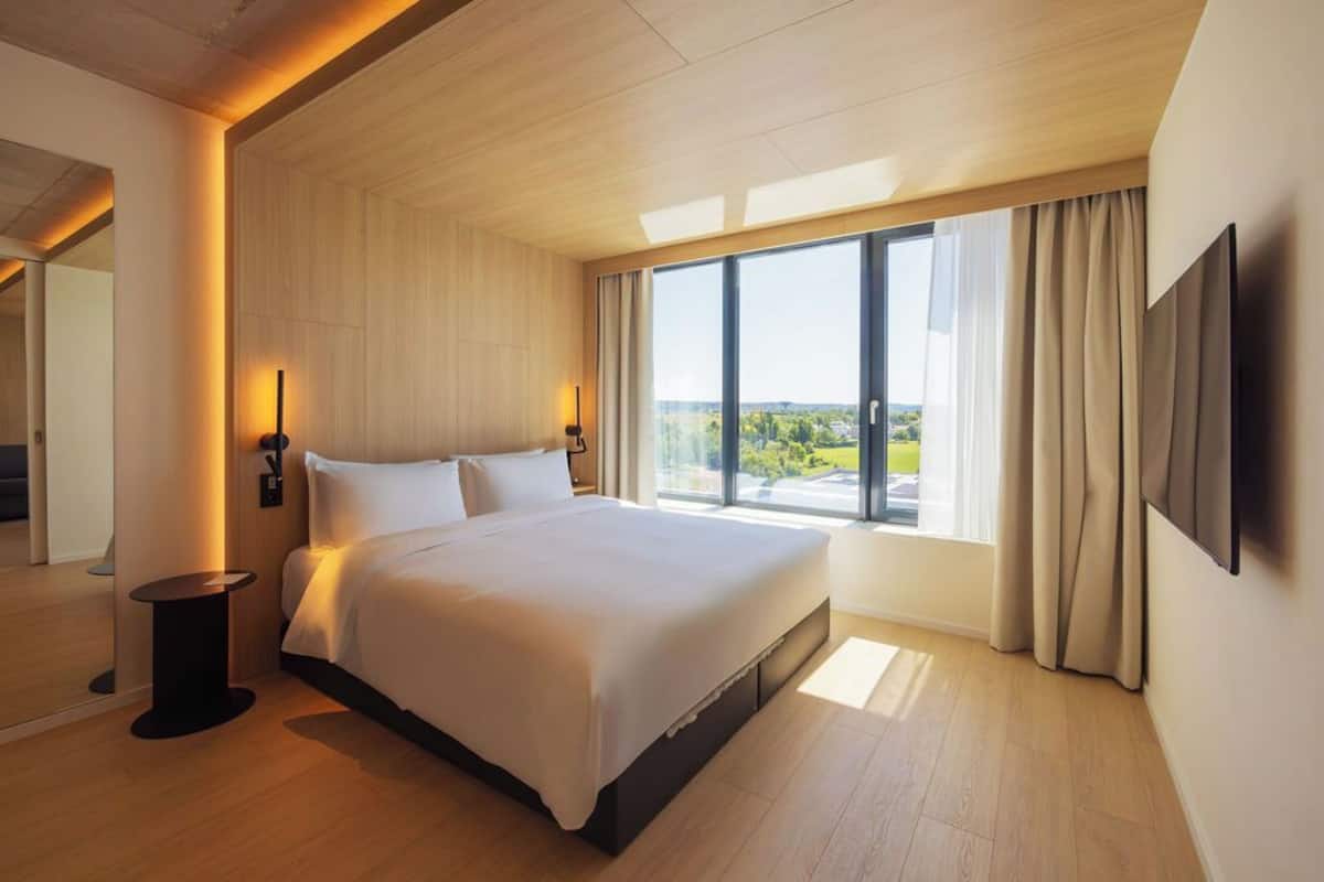 Una habitación de hotel moderna con una cama grande, un televisor montado en la pared y vistas al exterior.